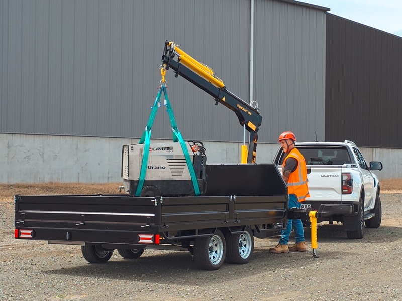 A tandem axle flatbed trailer with a Quicklift® 150-3 crane mounted at the front, stabiliser legs deployed, parked on a work site with the crane lifting a piece of machinery.