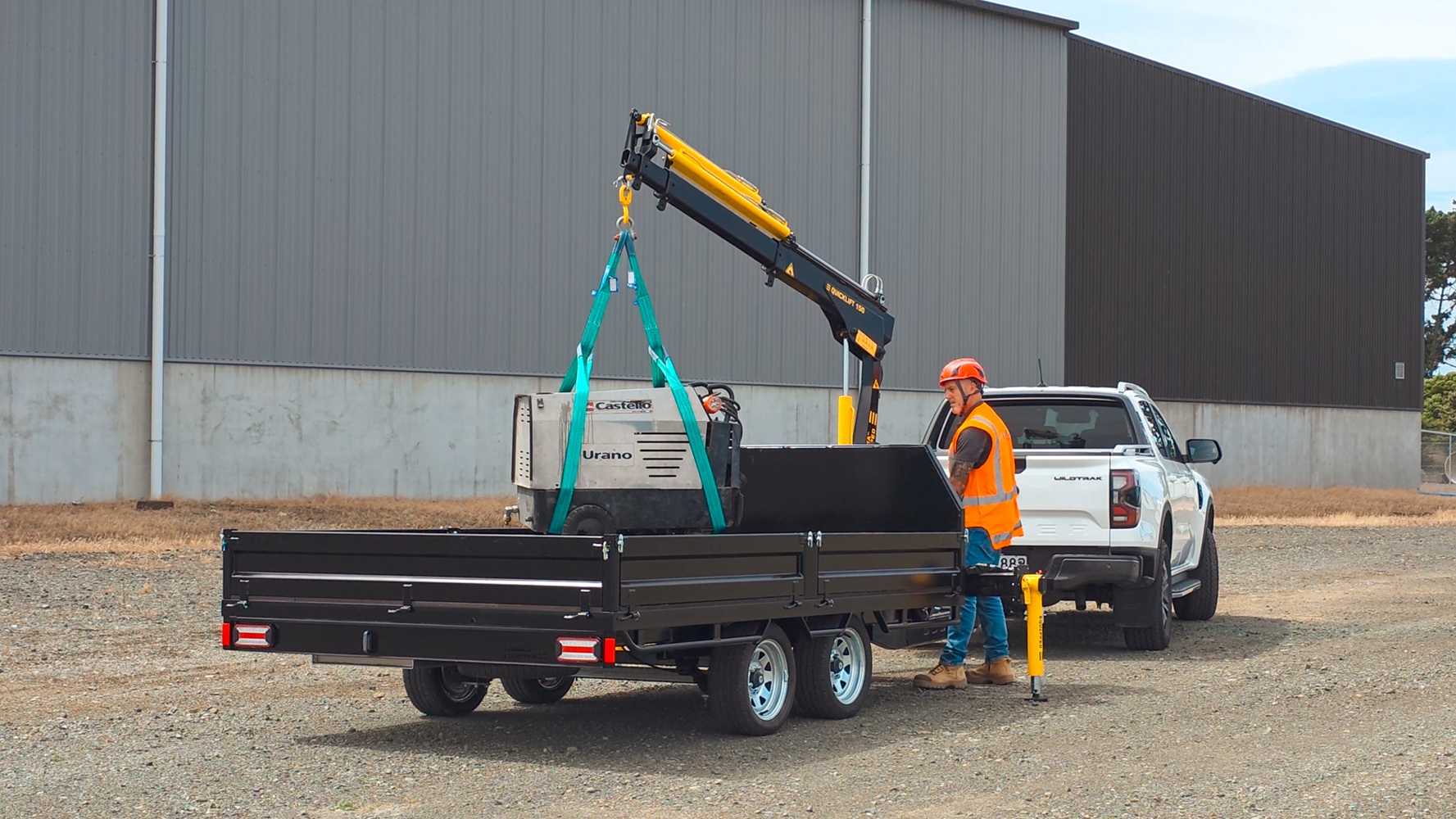 A tandem axle flatbed trailer with a Quicklift® 150-3 crane mounted at the front, stabiliser legs deployed, parked on a work site with the crane lifting a piece of machinery.