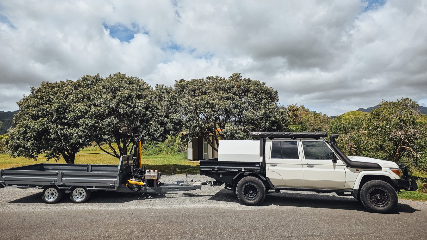 A tandem axle flatbed trailer with a Quicklift® 150-3 crane mounted at the front, stabiliser legs deployed, parked on a work site with the crane lifting a piece of machinery.