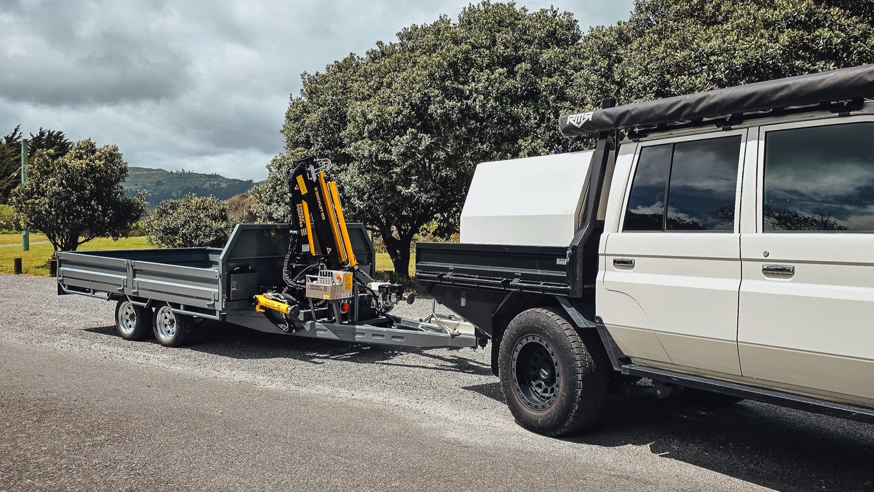 A tandem axle flatbed trailer with a Quicklift® 150-3 crane mounted at the front, stabiliser legs deployed, parked on a work site with the crane lifting a piece of machinery.