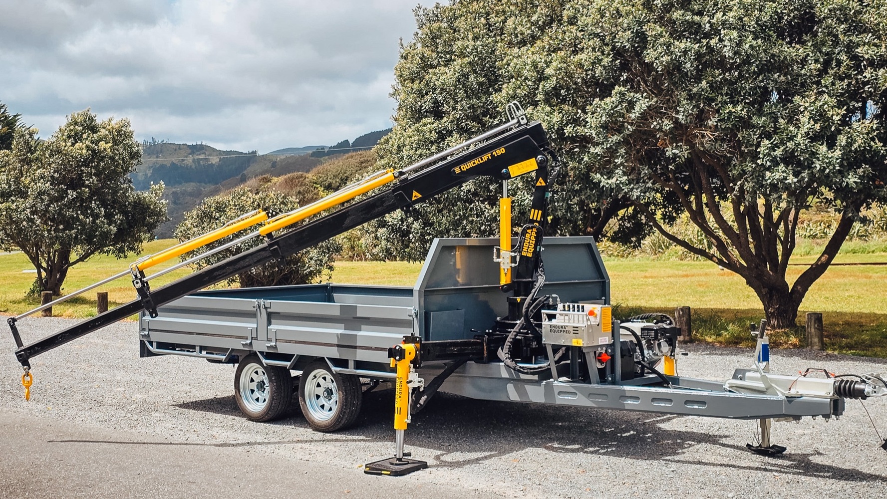 A tandem axle flatbed trailer with a Quicklift® 150-3 crane mounted at the front, stabiliser legs deployed, parked on a work site with the crane lifting a piece of machinery.