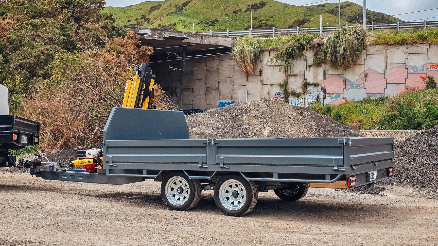 A tandem axle flatbed trailer with a Quicklift® 150-3 crane mounted at the front, stabiliser legs deployed, parked on a work site with the crane lifting a piece of machinery.