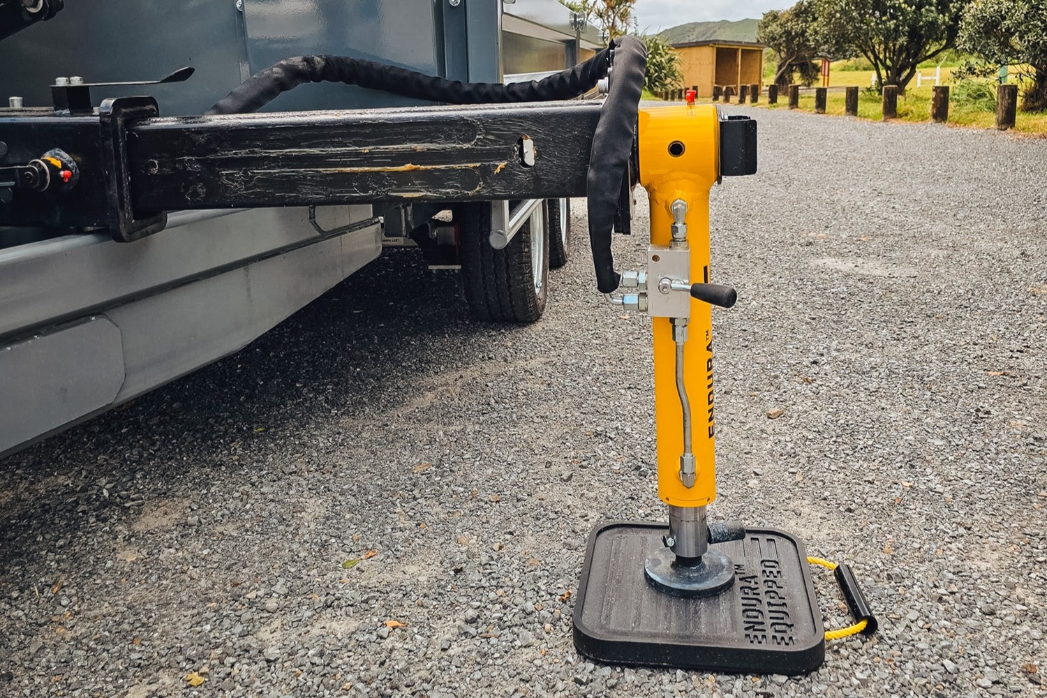 A tandem axle flatbed trailer with a Quicklift® 150-3 crane mounted at the front, stabiliser legs deployed, parked on a work site with the crane lifting a piece of machinery.